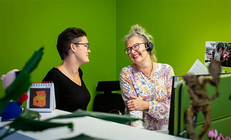 Två kvinnor sitter i en kontorsmiljö. Den ena har headset på sig och de ler mot varandra.
