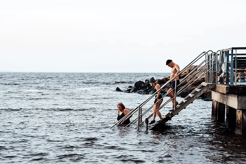 Tre personer går i havet från en trappa vid en brygga. 