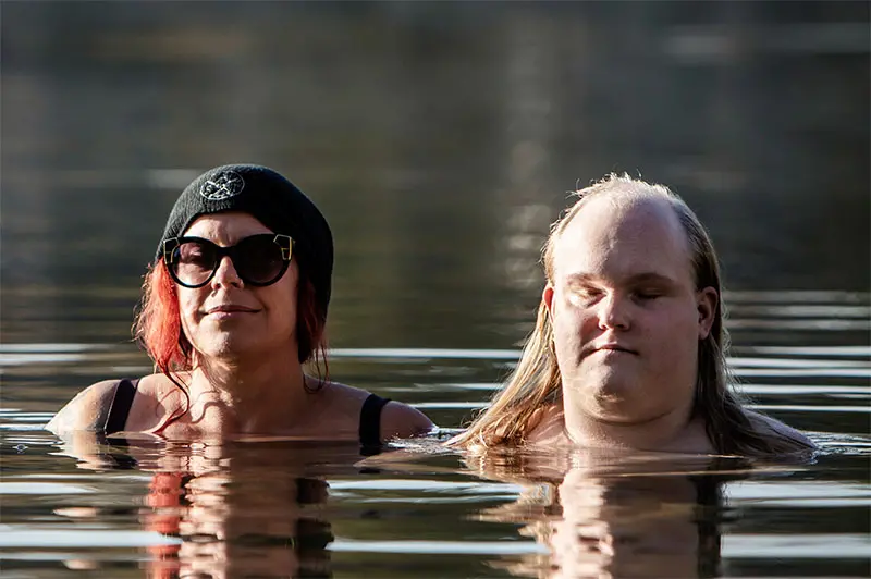 En närbild på kvinna och en man som badar i stilla, blankt vatten utomhus med vatten upp till halsen. De ser lugna ut. Kvinnan har mössa och solglasögon och ler. Mannen blundar.