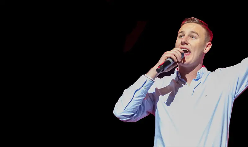 Alex Kron sjunger i en mikrofon som han håller i ena handen. Andra handen har han lyft och han tittar en aning uppåt. Han är klädd i skjorta.