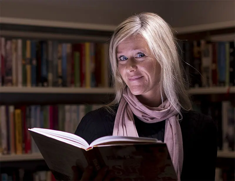 En kvinna tittar upp från en öppen bok som hon håller framför sig.