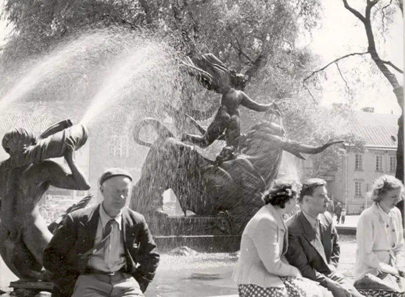 En arkivbild som visar Europa och tjuren med vattnet igång. I förgrunden sitter människor och vilar på fontänens kant. I bakgrunden syns träd runt fontänen.
