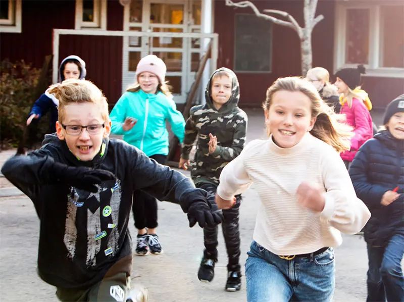 Glada barn springer mot kameran på en skolgård. Det är både tjejer och killar. Några har jacka och mössa, några är utan jacka.