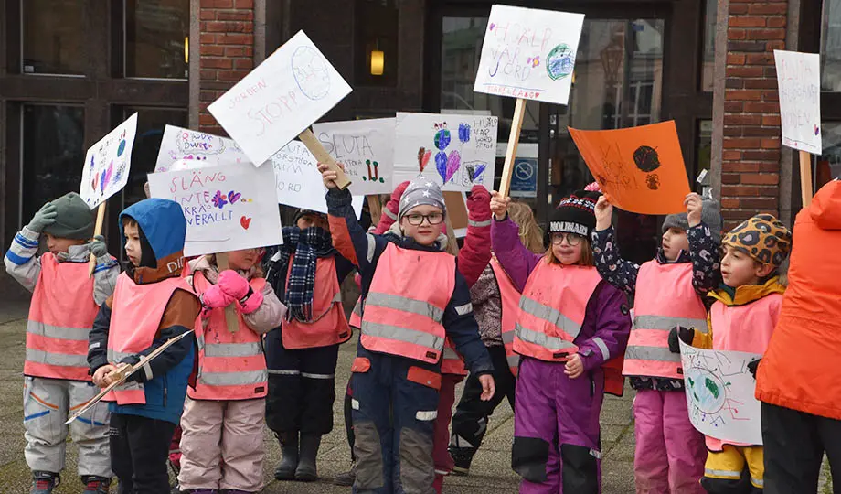 Tiotal barn i rosa reflexvästar håller upp skyltar med text och teckningar