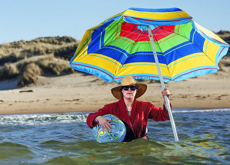 En kvinna sitter i havet med solhatt och parasoll.