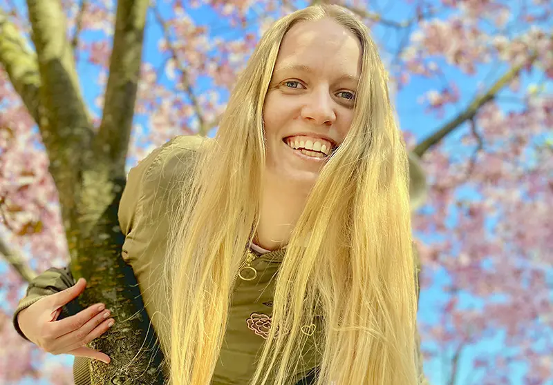 En kvinna med långt blont hår ler mot oss från ett körsbärsträd.