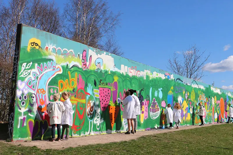 Workshop vid Halmstadväggen. Barn i olika åldrar målar graffiti på en lång vägg utomhus. 