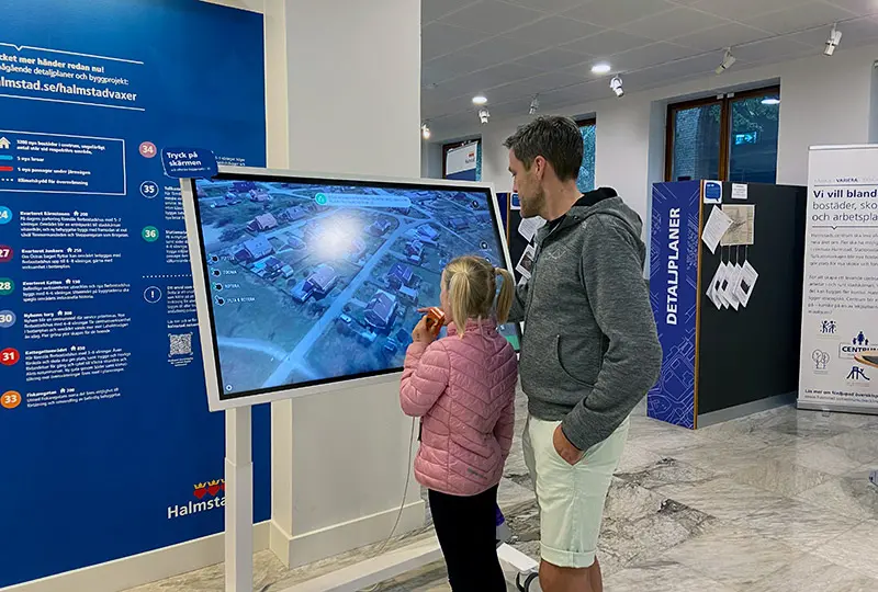 En pappa med sin dotter tittar på en skärm med 3D-visualisering av Halmstad. 